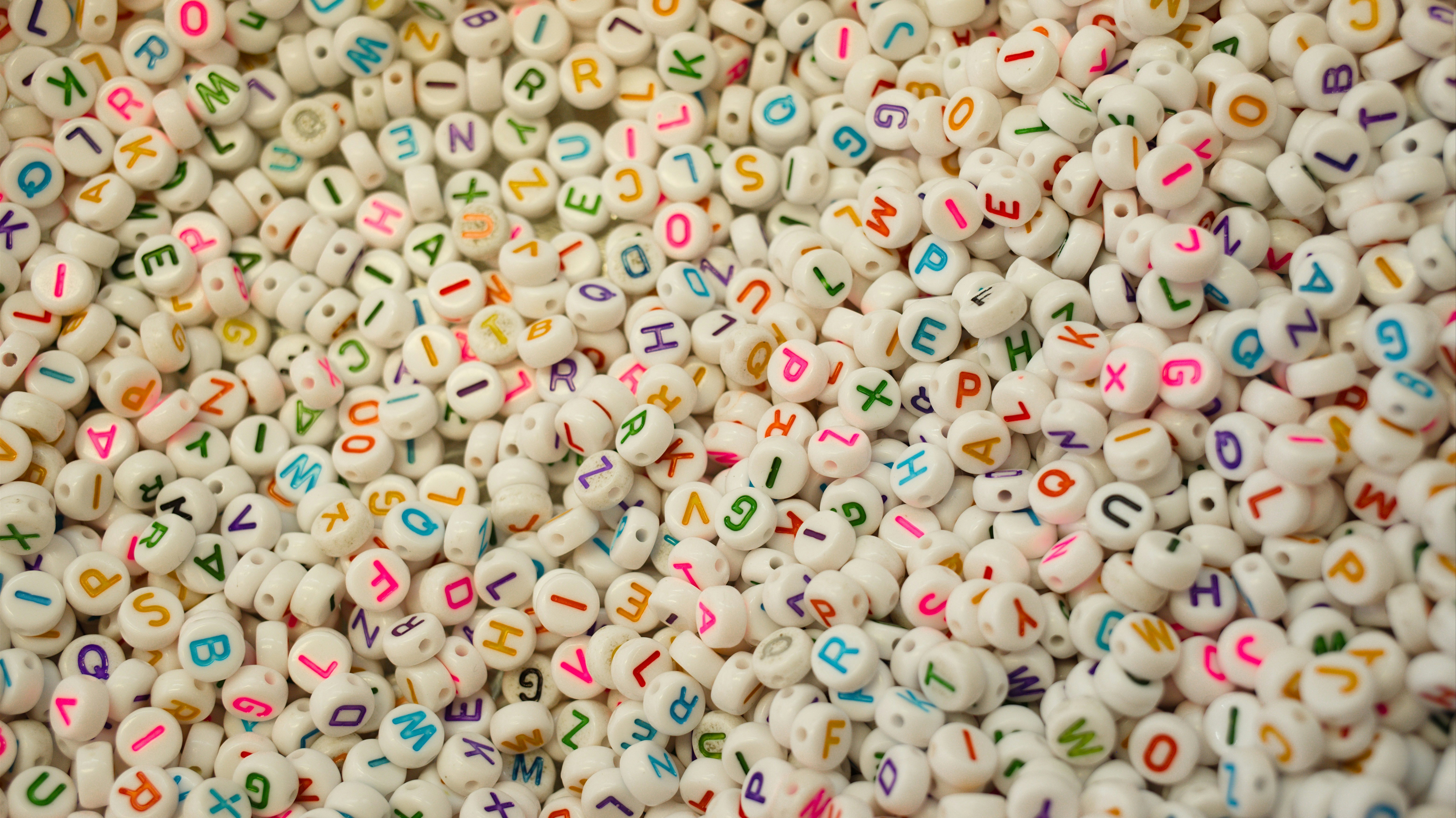 Une multitude de petites perles blanches ornées de lettres colorées est éparpillée en vrac, formant une mosaïque de caractères.